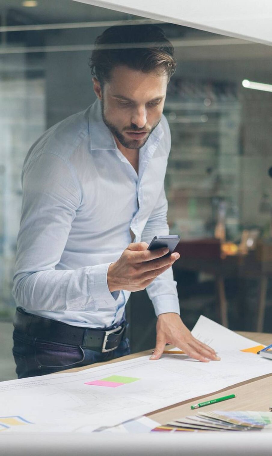 Man with beard wearing light blue shirt leans over a desk spread with papers and a model of a building, looking at a smartphone.
